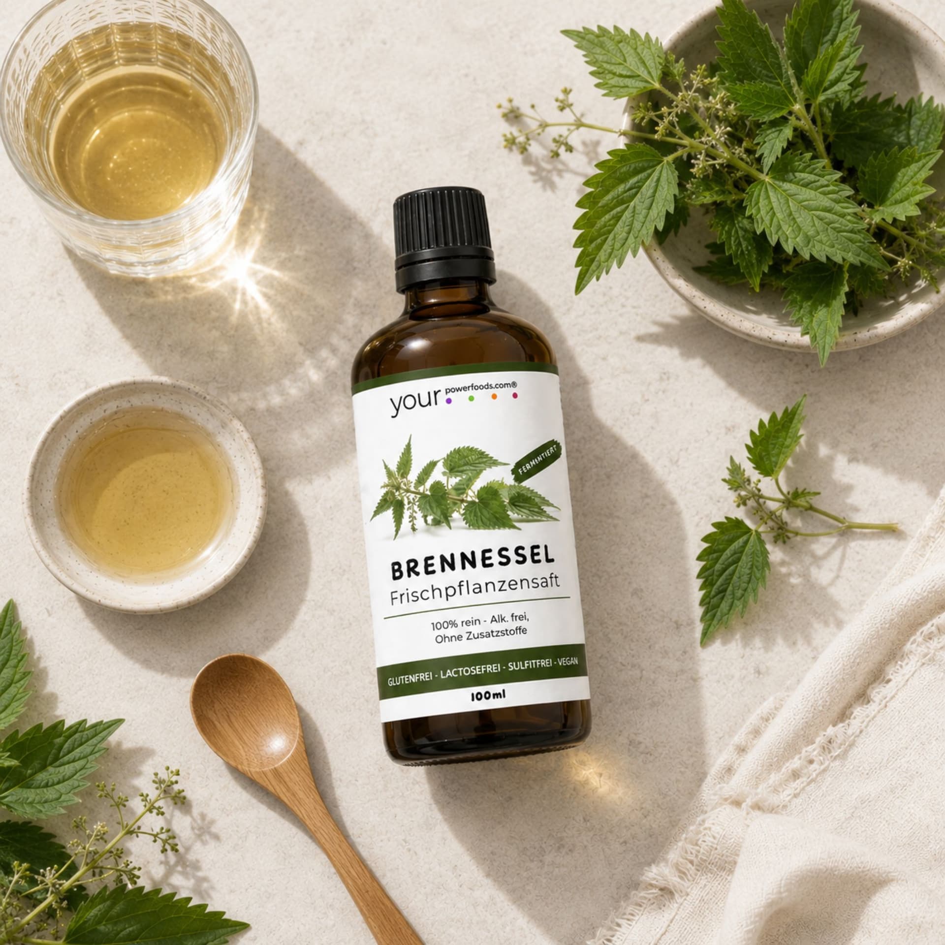 Brennessel - Elixier, fermentiert, 100 ml – braune Tropfflasche liegend, umgeben von frischen Brennesselblättern, Holzlöffel